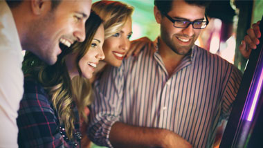 A man in a striped button down shirt and a man with a white shirt are enjoyng playing slots with two attractive women