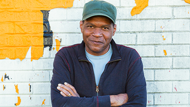 Robert Cray with his arms crossed in front of a painted brick wall.