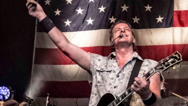 Ted Nugent holding guitar and hat with American flag background