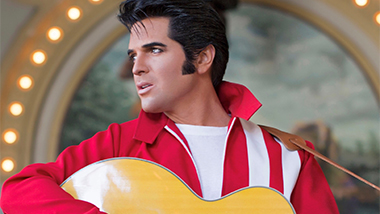 Dean Z, The Ultimate Elvis, in a red jacket with an acoustic guitar looking off camera to the left.